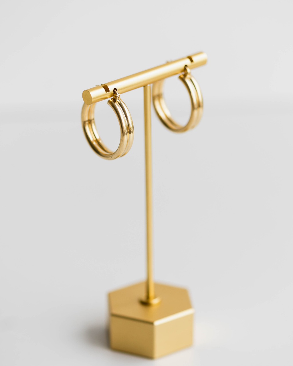 Gold hoop earrings on a gold T-shaped stand with a hexagonal base against a light gray background