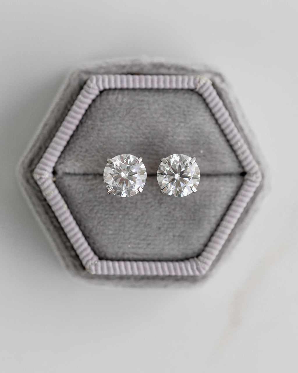 Pair of diamond stud earrings on a hexagonal gray fabric stand against a light gray background