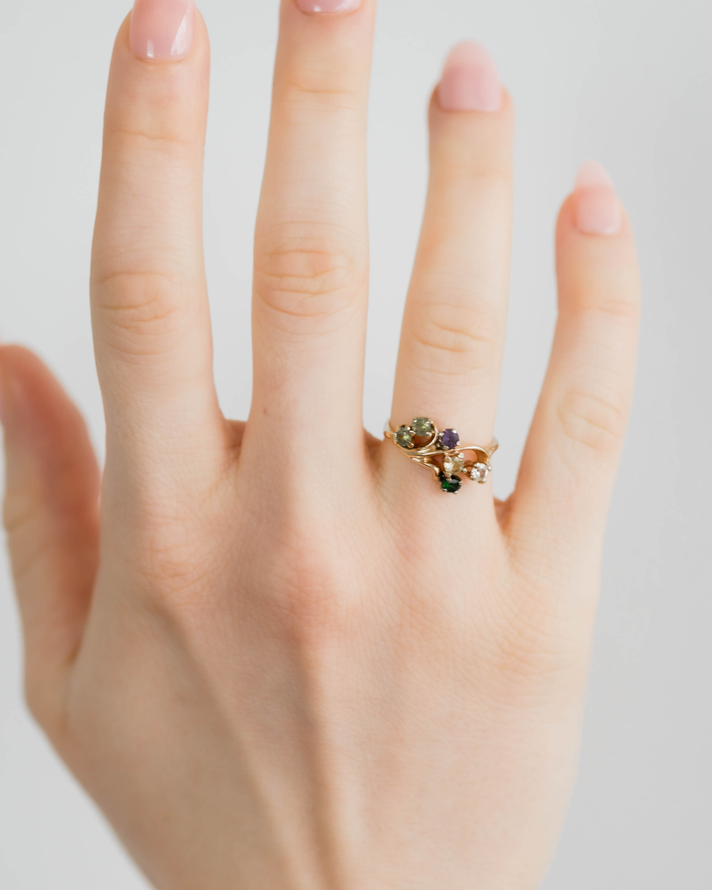 Hand wearing a gold ring with green and purple gemstones on a light background